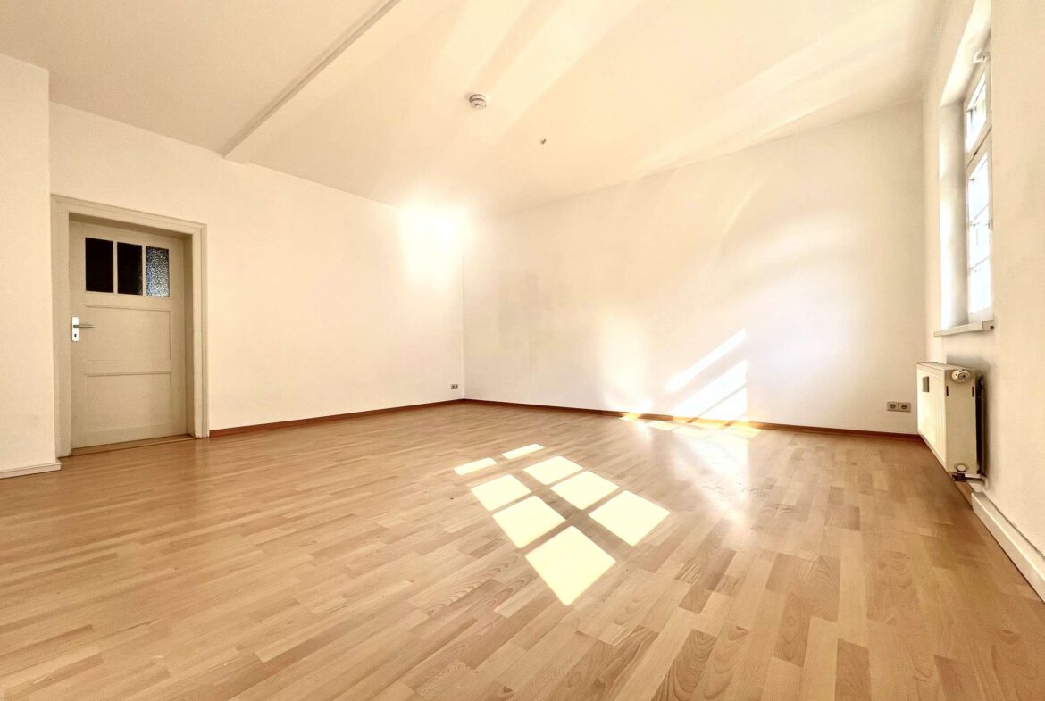 Bright empty room with light wood floor, white walls, a door on the left, and sunlight patterns on the floor.