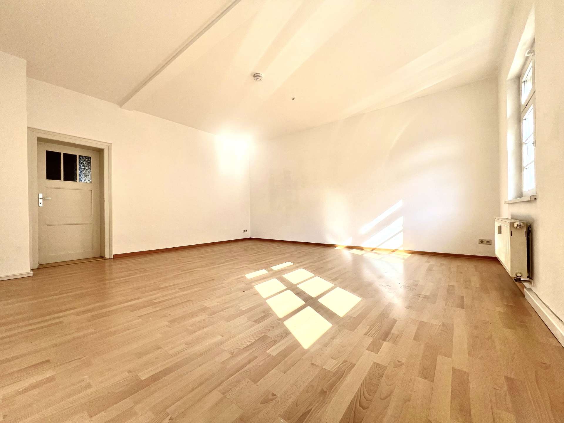 Bright empty room with light wood floor, white walls, a door on the left, and sunlight patterns on the floor.