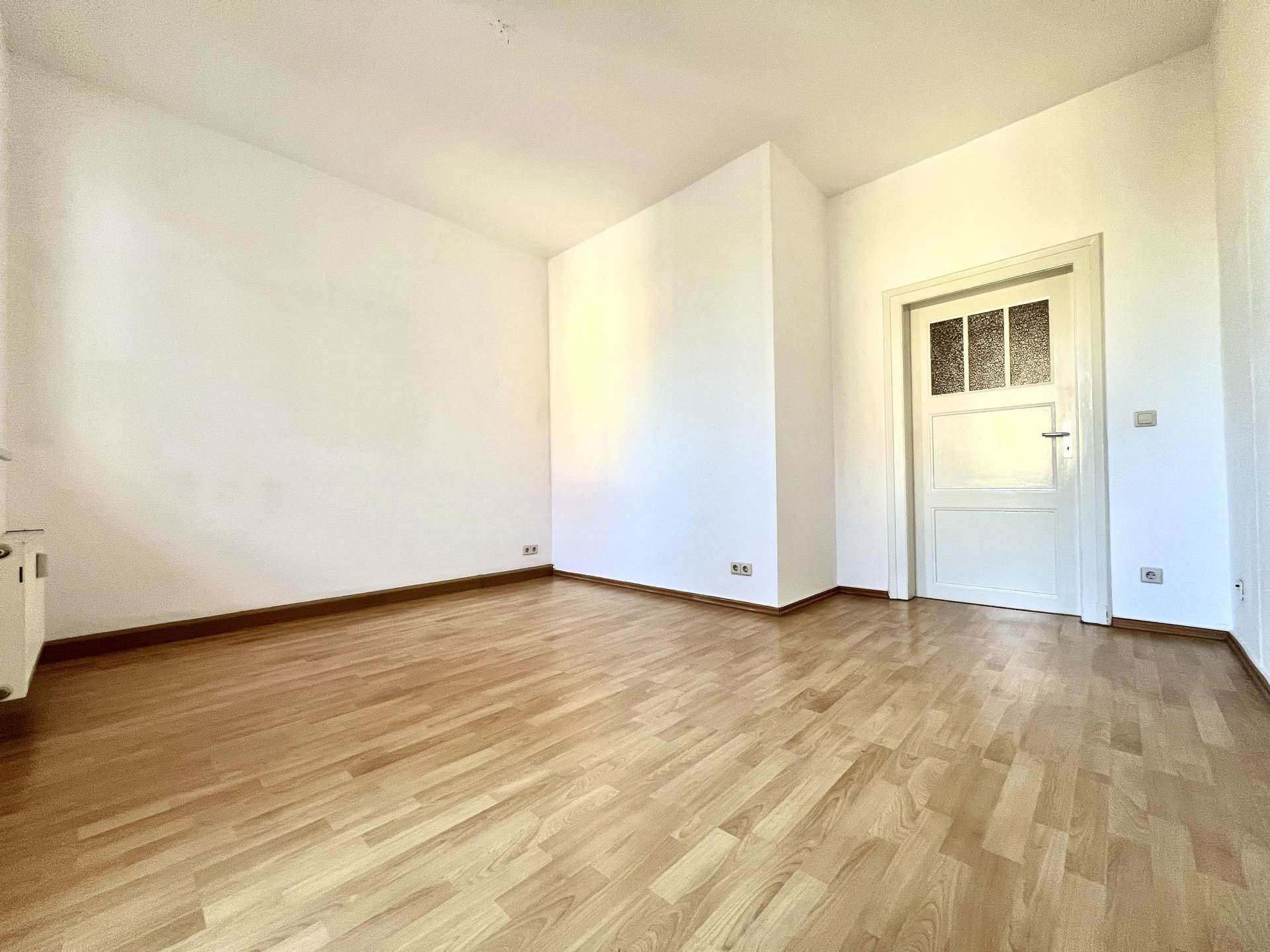 Empty bright room with light wood flooring, white walls, and a white door on the right side. narrow corner wall creates a diagonal light/shadow line.