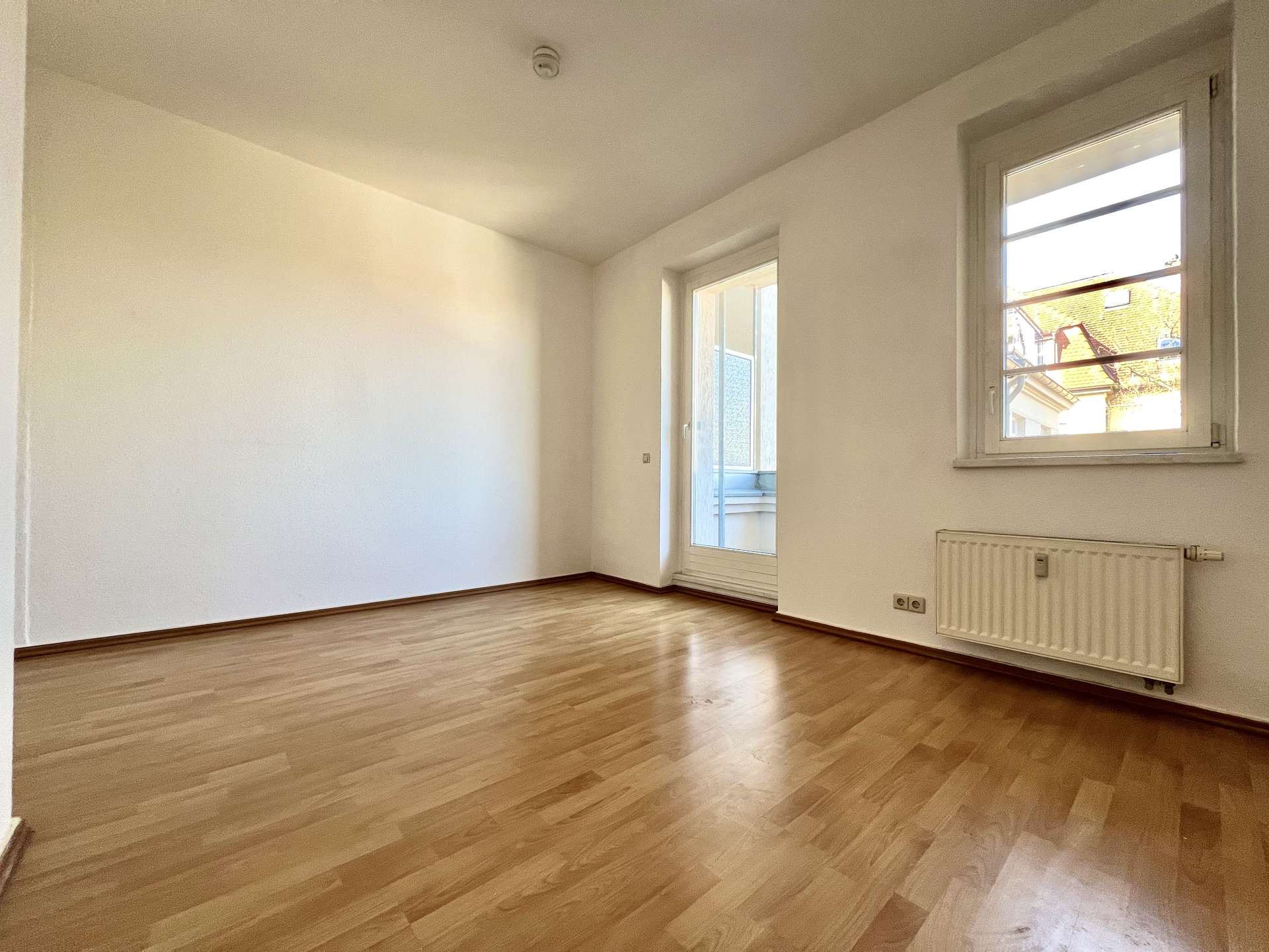 Bright empty room with light wood flooring, white walls, a large window on the right, and a radiator below it.