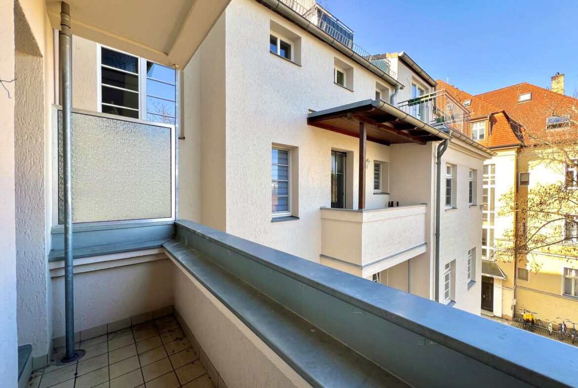 Balcony of a cream-colored apartment building with tiled floor and metal railing, blue sky above, neighboring red-tiled buildings in distance