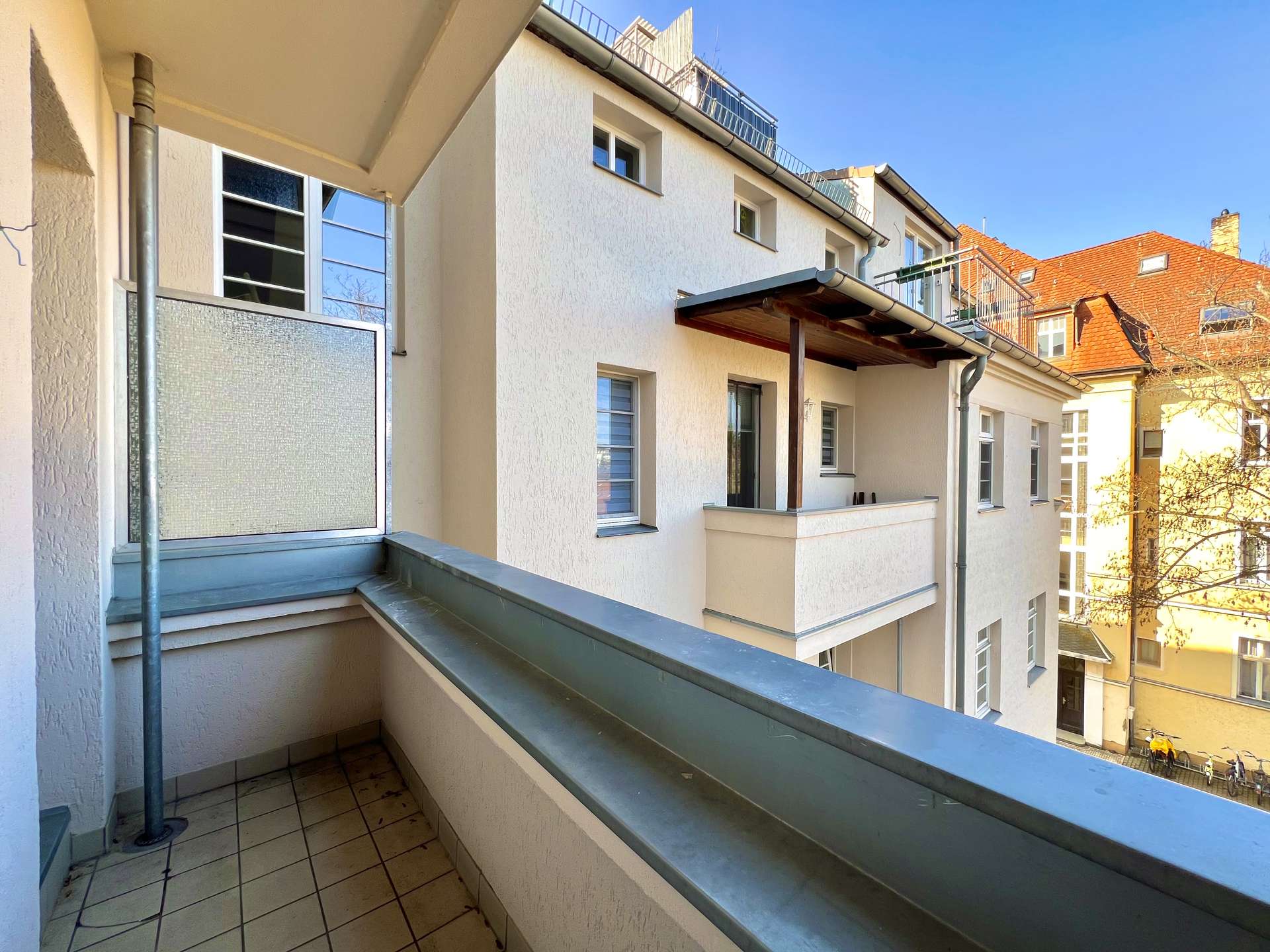 Balcony of a cream-colored apartment building with tiled floor and metal railing, blue sky above, neighboring red-tiled buildings in distance