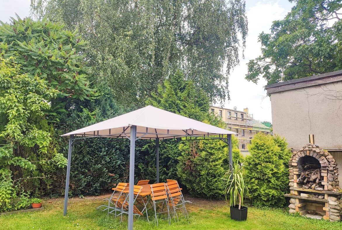 Backyard with a metal gazebo, orange folding chairs around a table, and lush green trees surrounding the space, plus a brick wood-fired oven on the right.