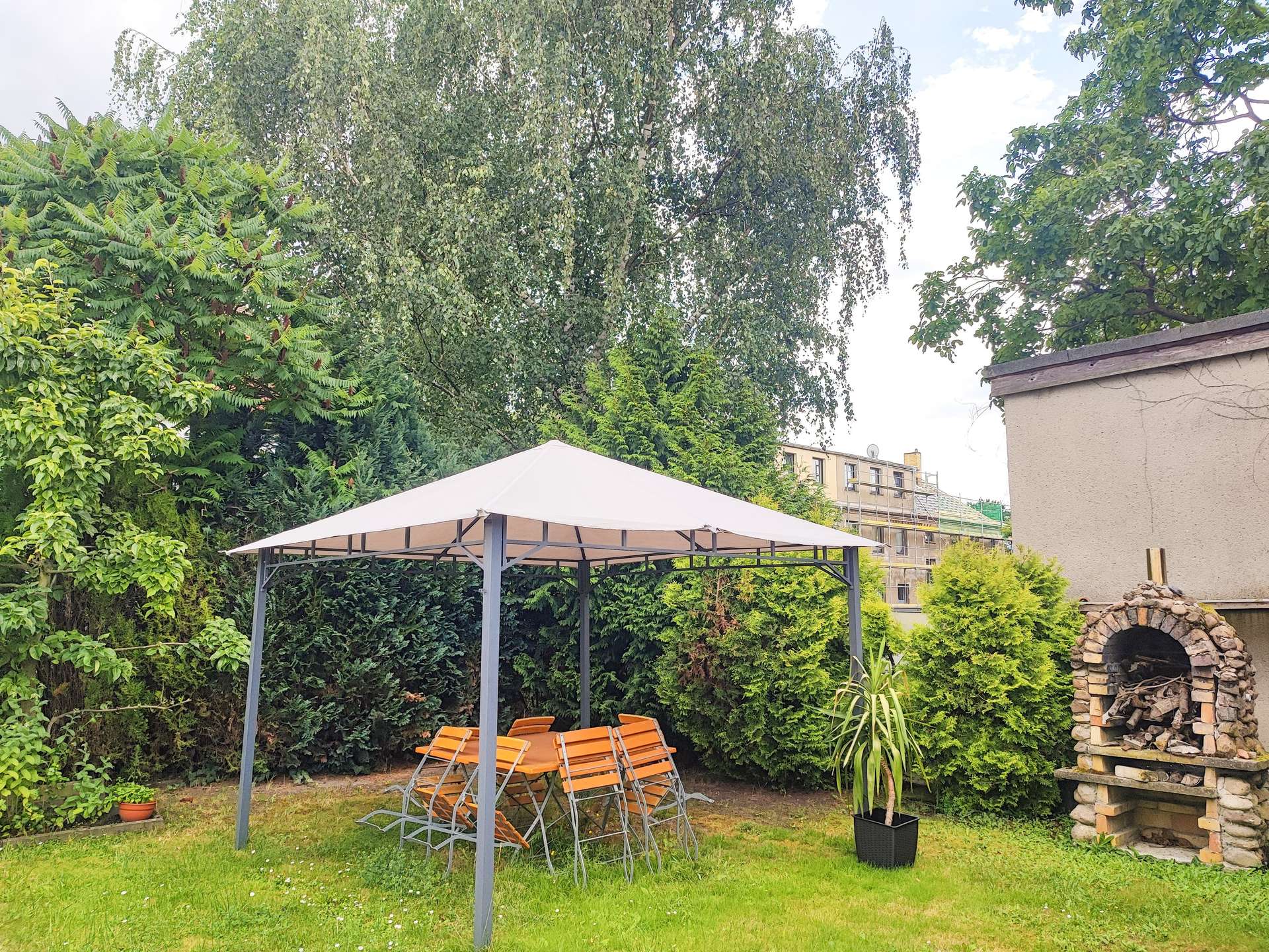 Backyard with a metal gazebo, orange folding chairs around a table, and lush green trees surrounding the space, plus a brick wood-fired oven on the right.