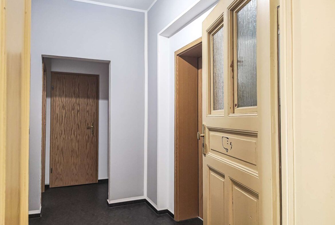 Narrow hallway with beige front door featuring frosted glass panels on the right and wooden doors along gray walls on the left and back.