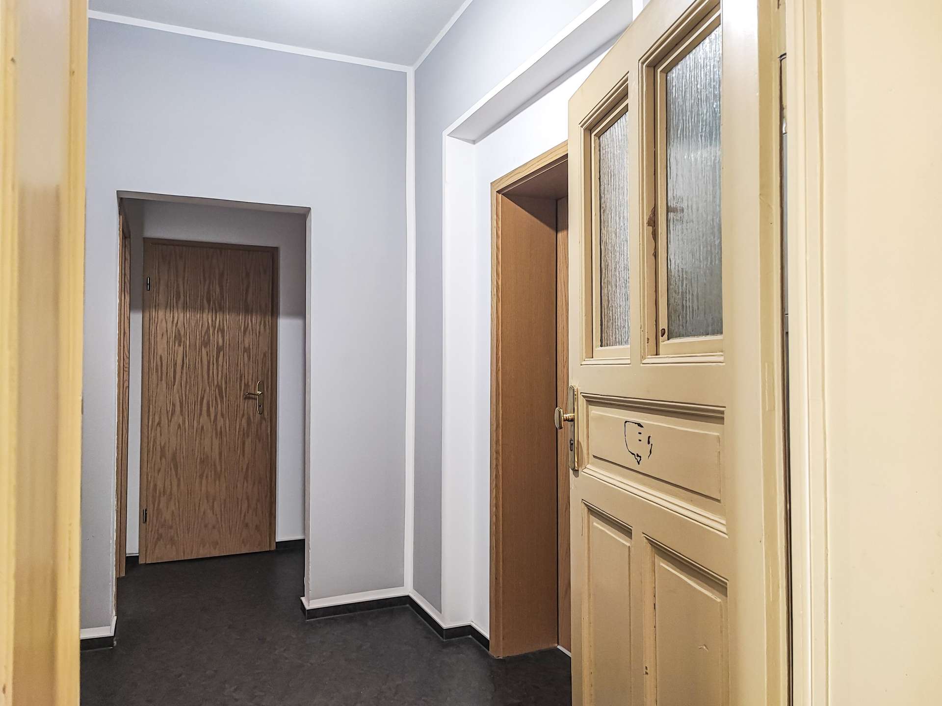 Narrow hallway with beige front door featuring frosted glass panels on the right and wooden doors along gray walls on the left and back.