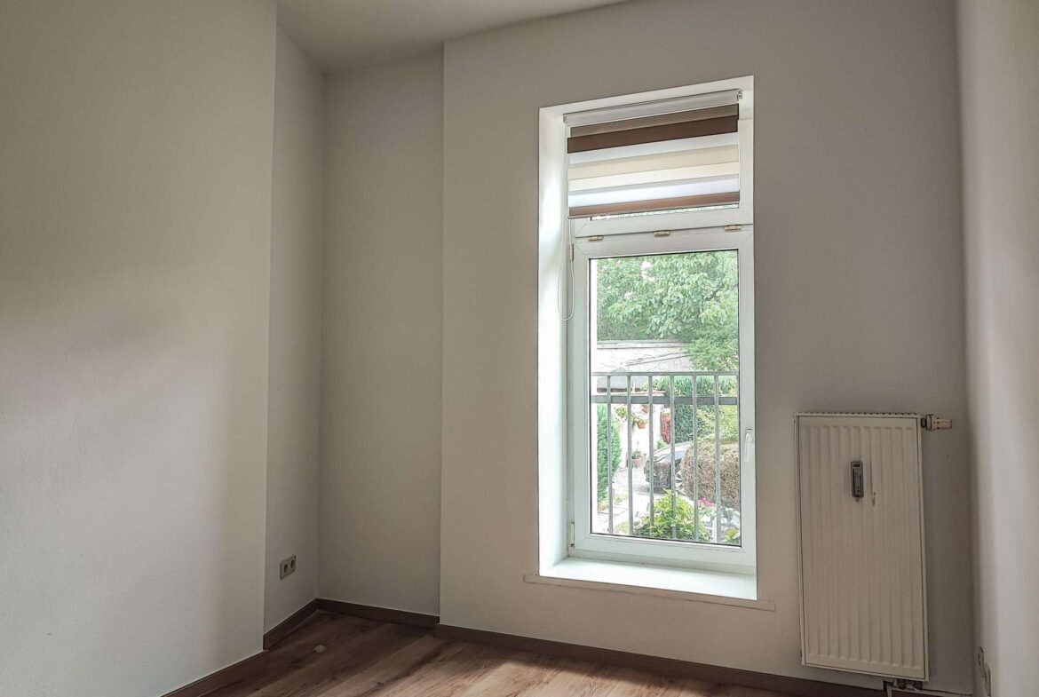 Empty small room with light gray walls, wood floor, and a single window with striped blinds; radiator on the right wall.