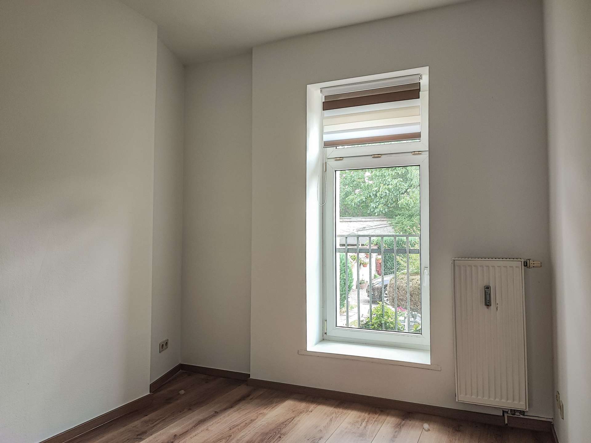 Empty small room with light gray walls, wood floor, and a single window with striped blinds; radiator on the right wall.