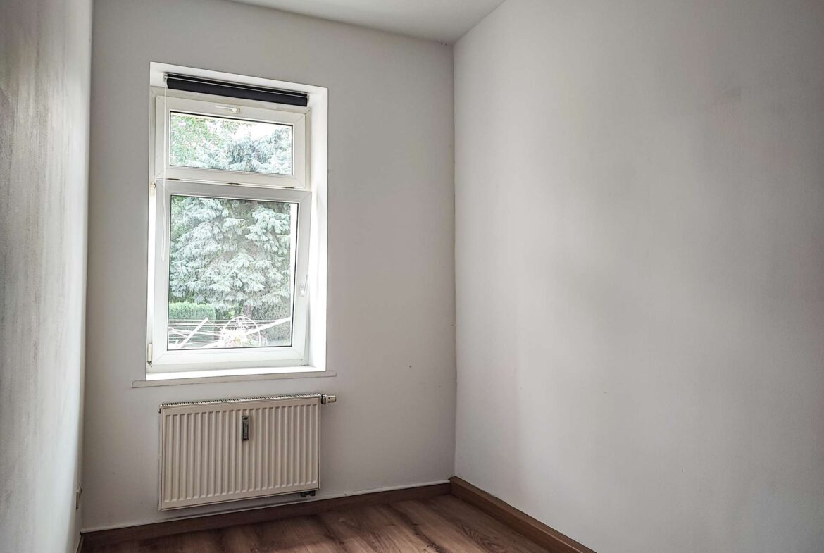 Empty small room with white walls, a window letting in light, and a white radiator beneath the window; wood-looking flooring visible. Outside, green trees are seen through the window.
