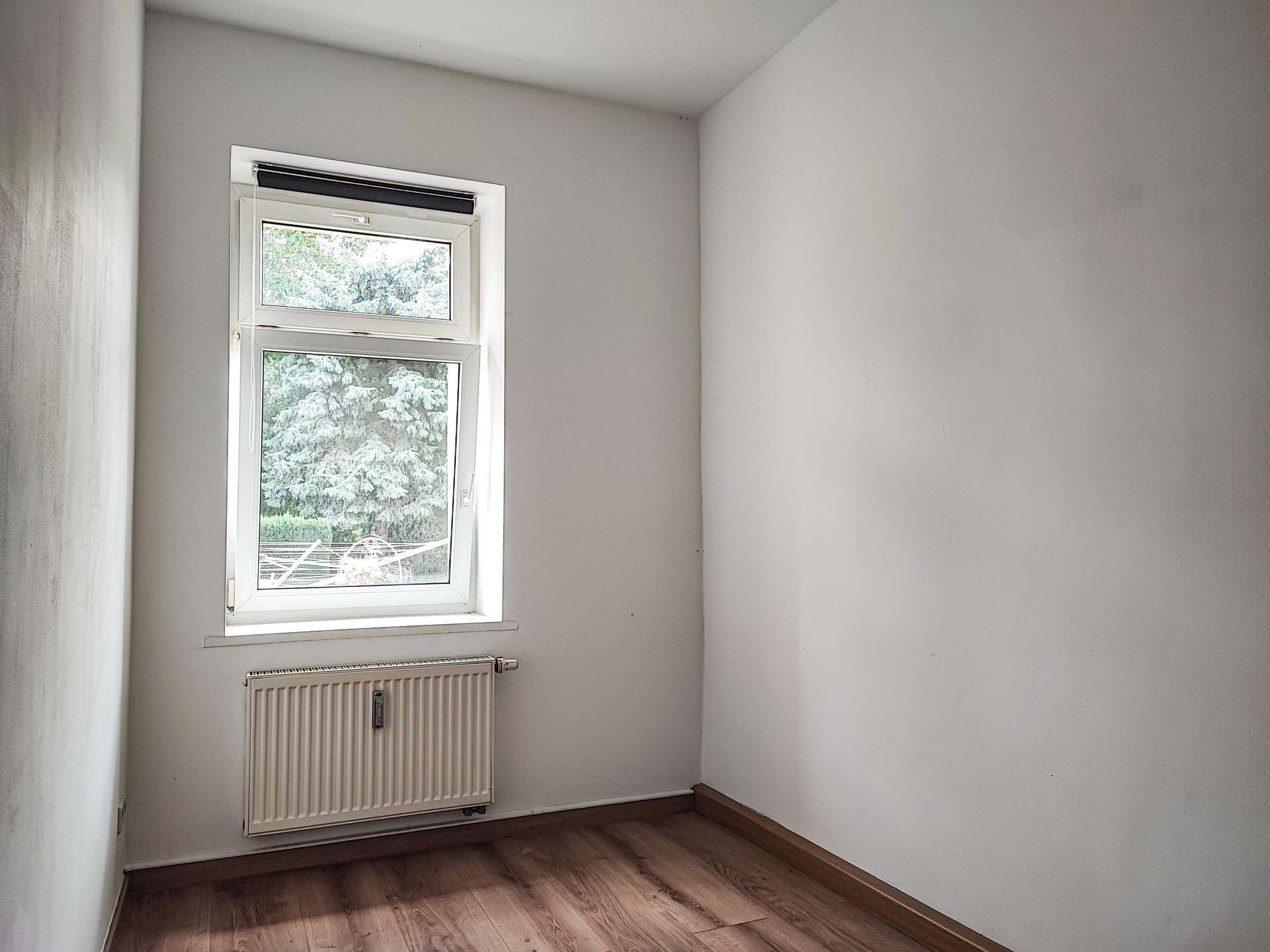 Empty small room with white walls, a window letting in light, and a white radiator beneath the window; wood-looking flooring visible. Outside, green trees are seen through the window.