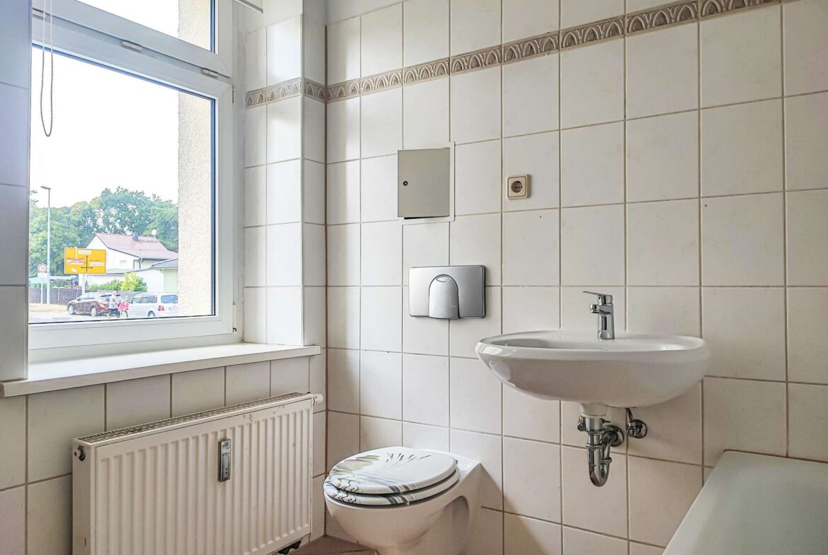 Bright tiled bathroom interior with a large window, radiator, wall-mounted fixtures, toilet, and sink visible.