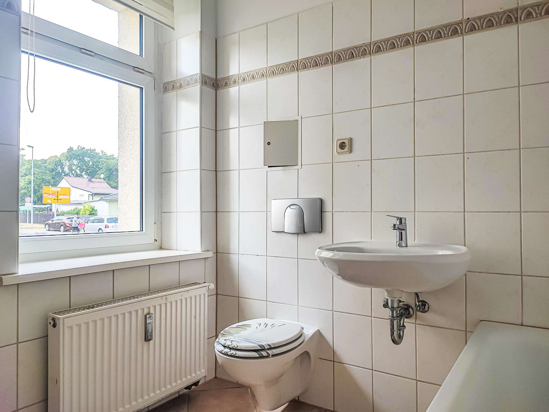 Bright tiled bathroom interior with a large window, radiator, wall-mounted fixtures, toilet, and sink visible.