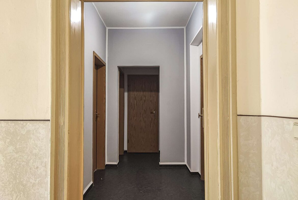 Long, dim hallway viewed from an open doorway, with multiple wooden doors along gray-walled corridors and beige trim.