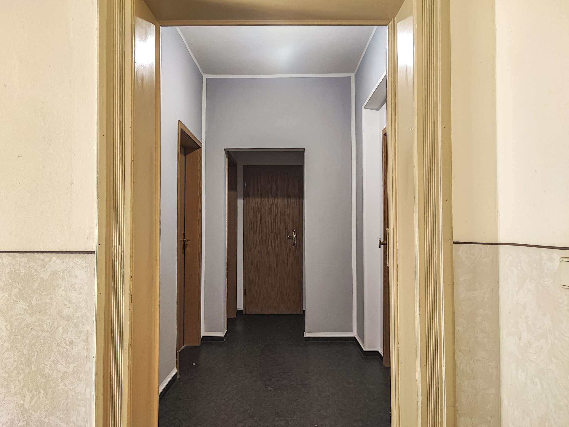 Long, dim hallway viewed from an open doorway, with multiple wooden doors along gray-walled corridors and beige trim.
