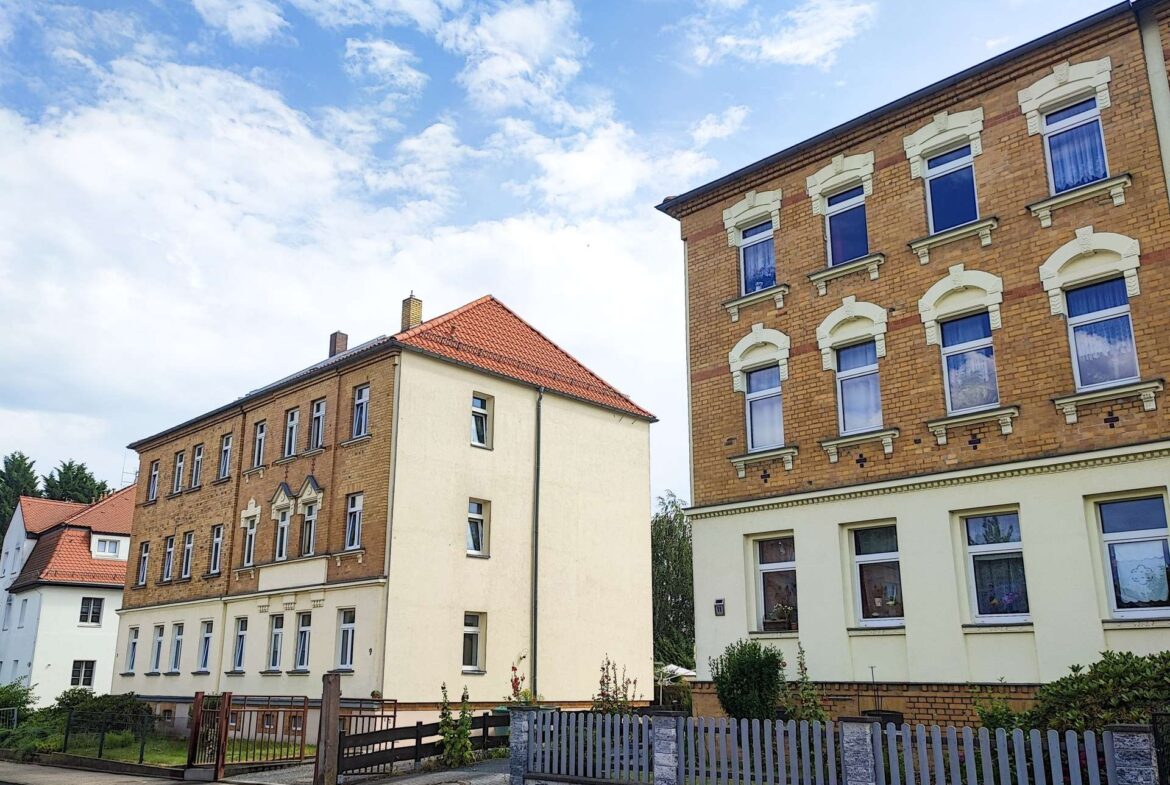 Red-brick apartment building with white decorative window arches on a sunny day, next to a beige multi-story building; wooden posts and a low fence in the foreground.