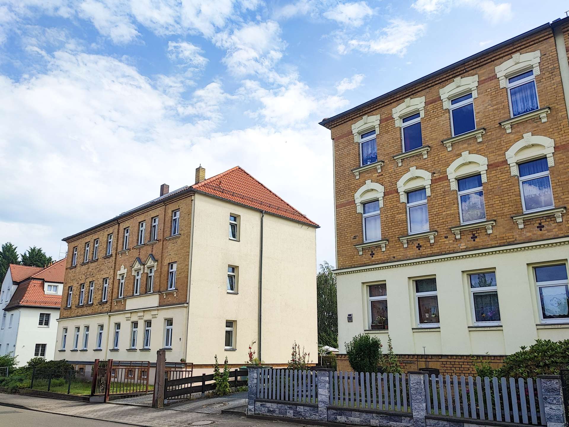 Red-brick apartment building with white decorative window arches on a sunny day, next to a beige multi-story building; wooden posts and a low fence in the foreground.