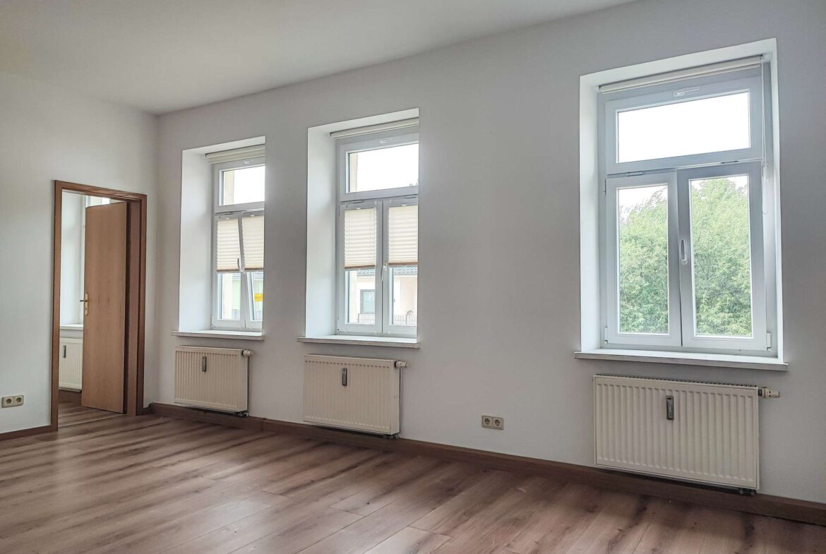 Empty room with light wood floors, three large vertical windows with blinds and radiators beneath, and an open wooden door on the left.