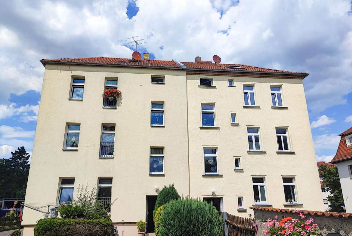 Front view of a beige multi-story apartment building with red-tiled roof and satellite dishes under a blue sky with clouds, windows visible.