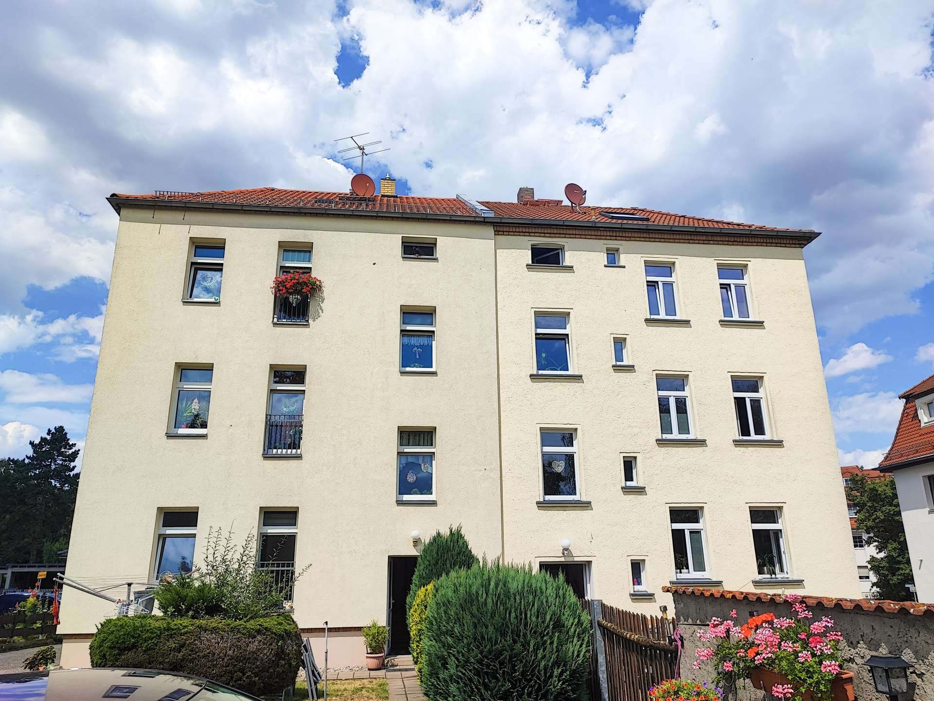 Front view of a beige multi-story apartment building with red-tiled roof and satellite dishes under a blue sky with clouds, windows visible.