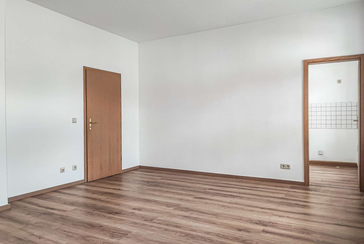 Empty room with light wood floors, white walls, and a wooden door on the left; a tall mirror with a wooden frame is mounted on the right wall showing tiled area.