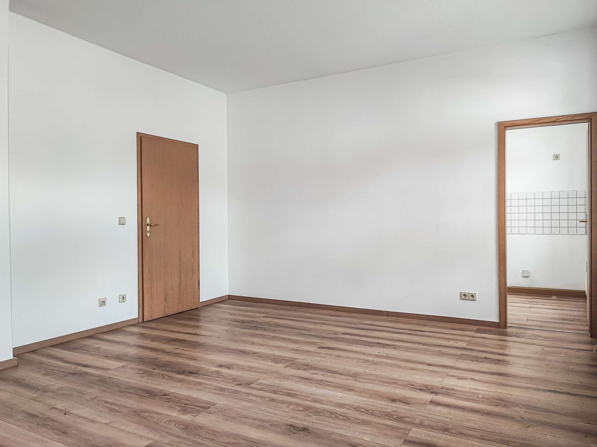 Empty room with light wood floors, white walls, and a wooden door on the left; a tall mirror with a wooden frame is mounted on the right wall showing tiled area.
