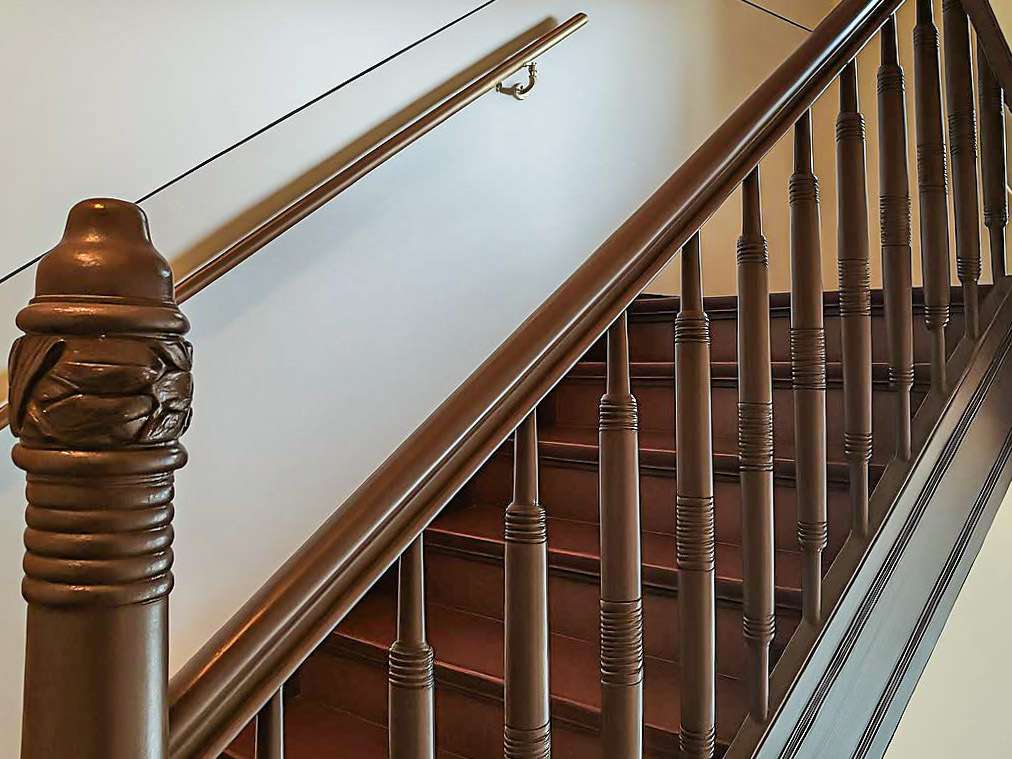 Wooden staircase with a carved newel post and brown handrail seen from the bottom corner edge of a hallway.