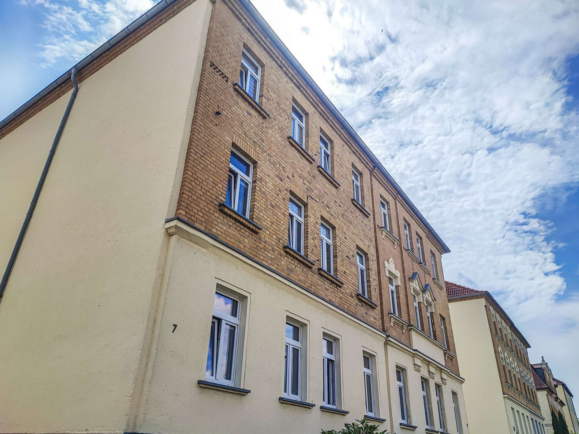 Angle view of a brick apartment building with multiple white-framed windows against a blue, cloudy sky.