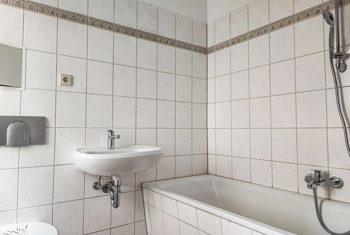 White tiled bathroom with a wall-mounted sink, chrome faucet, and a bathtub along the right; shower head on the wall and a towel dispenser on the left.