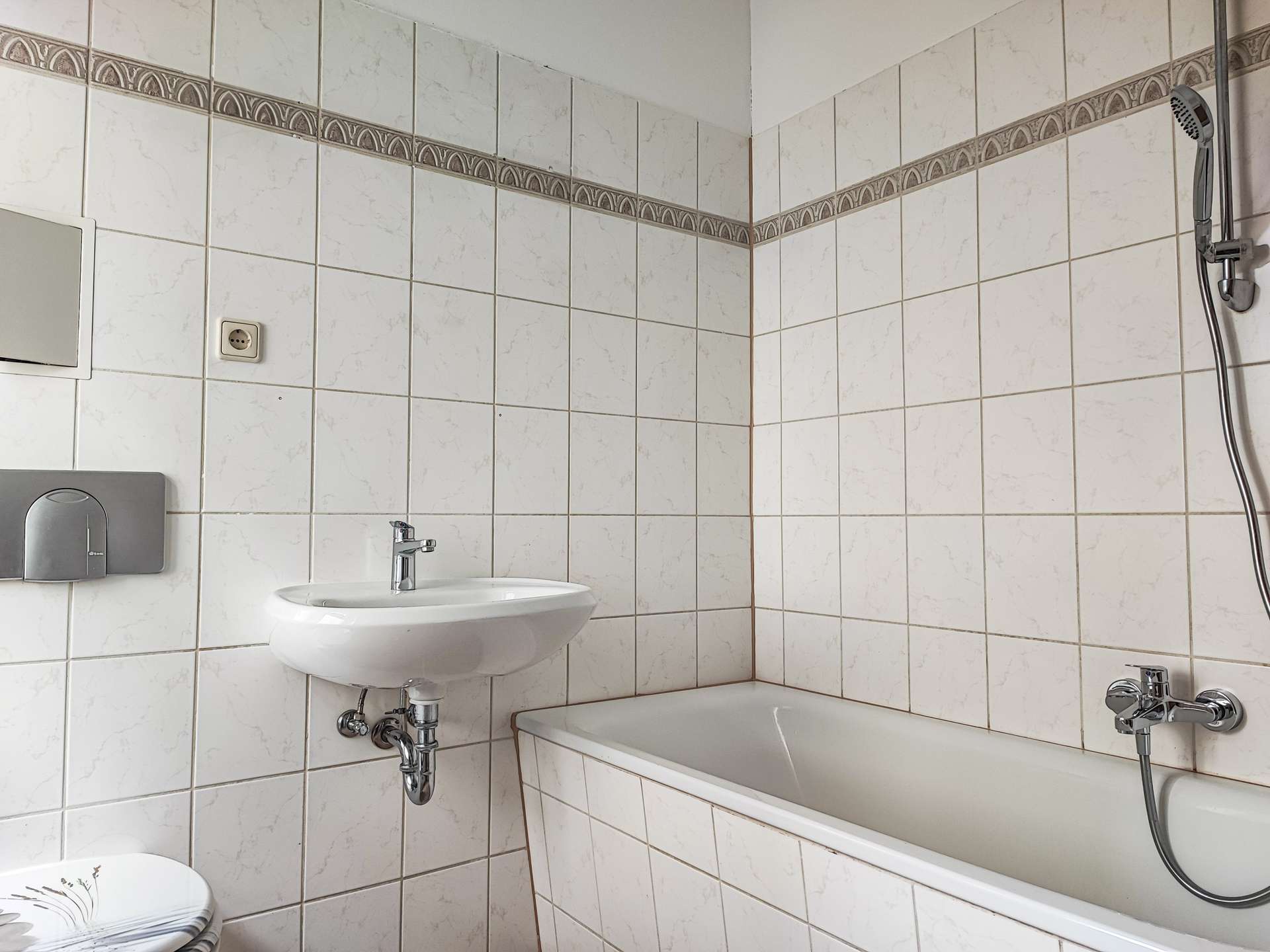 White tiled bathroom with a wall-mounted sink, chrome faucet, and a bathtub along the right; shower head on the wall and a towel dispenser on the left.