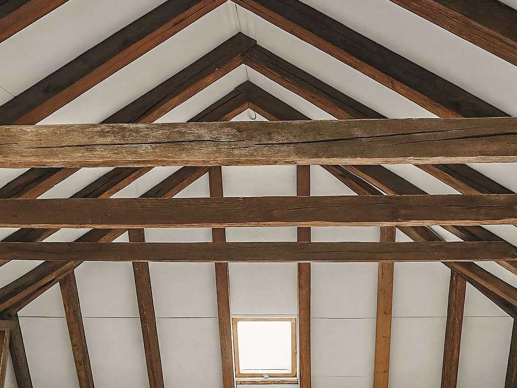 Timber-framed attic ceiling with exposed brown trusses forming a triangular peak, and a small window at the far end.