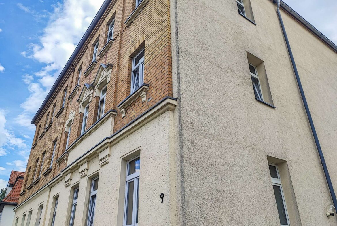 Corner view of a brick apartment building with beige stucco side and white-framed windows against a blue sky with clouds, number 9 on wall near door.