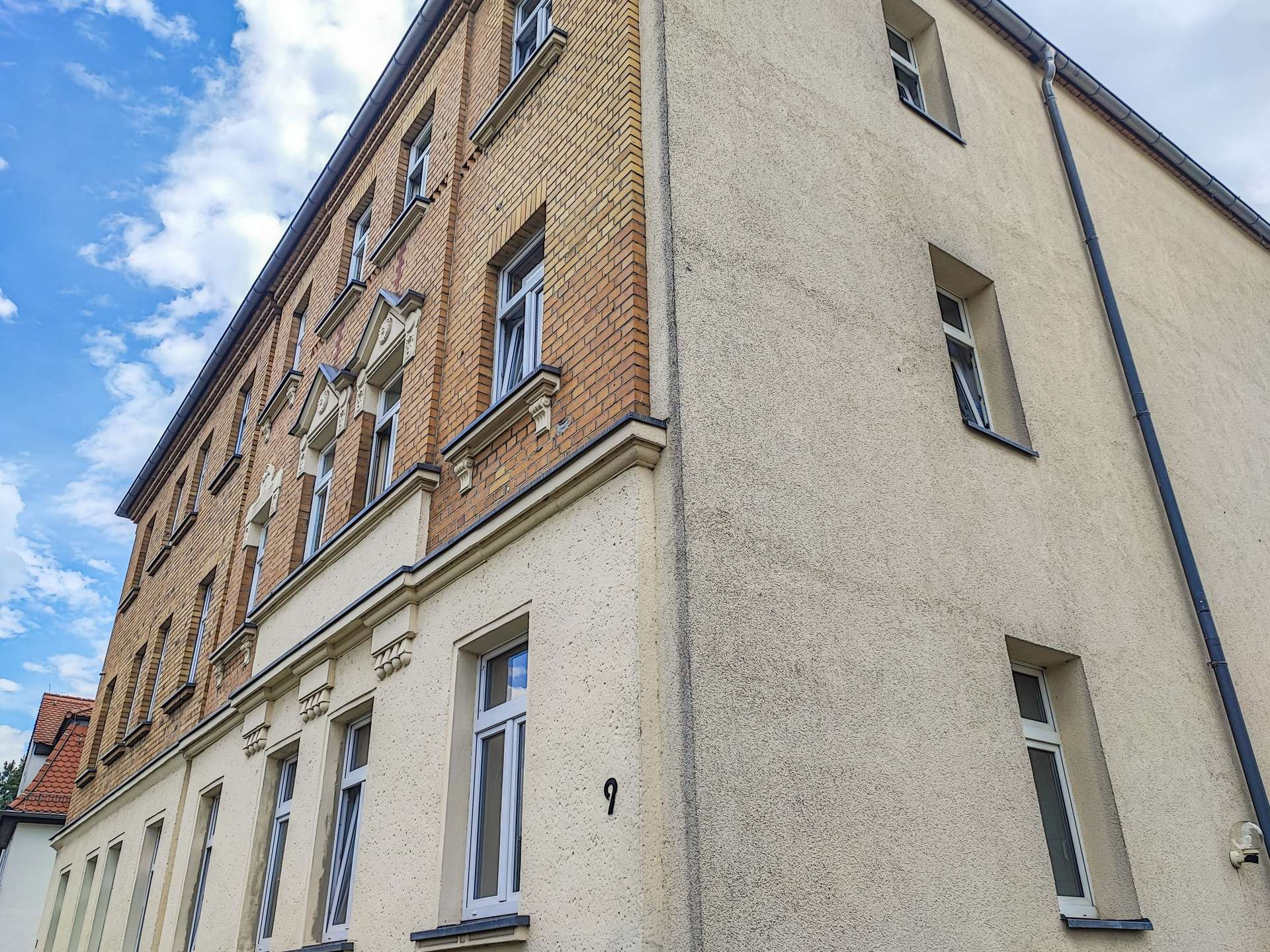 Corner view of a brick apartment building with beige stucco side and white-framed windows against a blue sky with clouds, number 9 on wall near door.