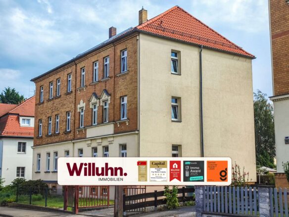 Facade of a multi-story brick apartment building with a red tiled roof and a large Willuhn Immobilien sign in the foreground, displaying award logos.