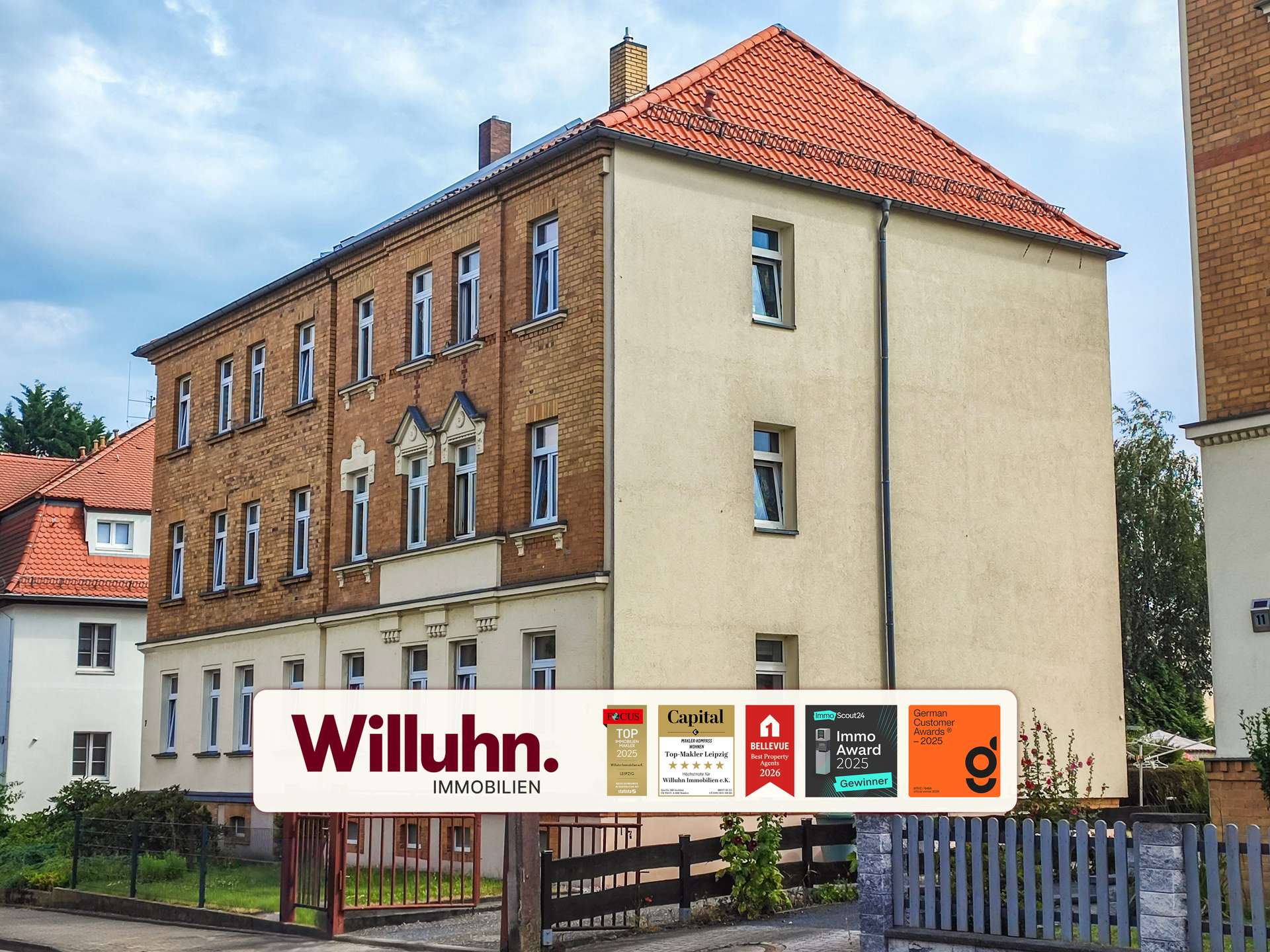 Facade of a multi-story brick apartment building with a red tiled roof and a large Willuhn Immobilien sign in the foreground, displaying award logos.