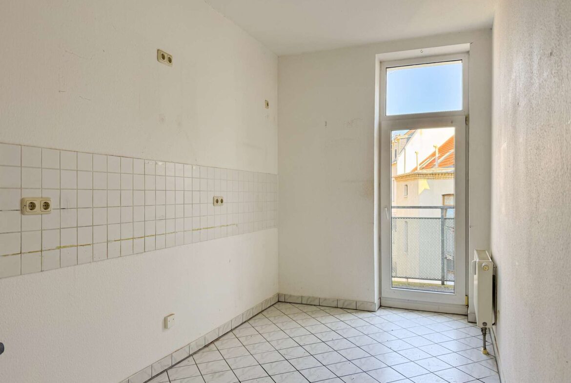 Empty kitchen corner with white walls and tiled backsplash, a balcony door letting in daylight, and a radiator to the right.