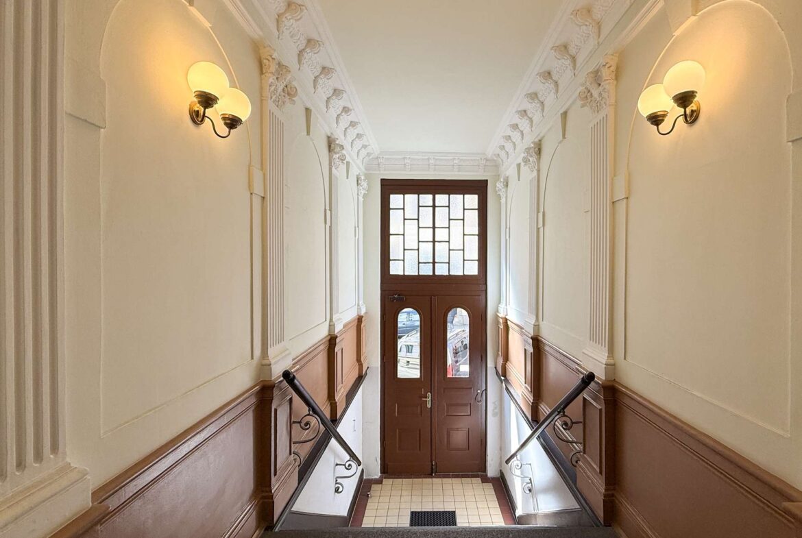 Elegant vintage hallway with ornate plaster molding, white walls, and warm wall lamps leading to a wooden door with frosted glass panes at the bottom of the stairs.