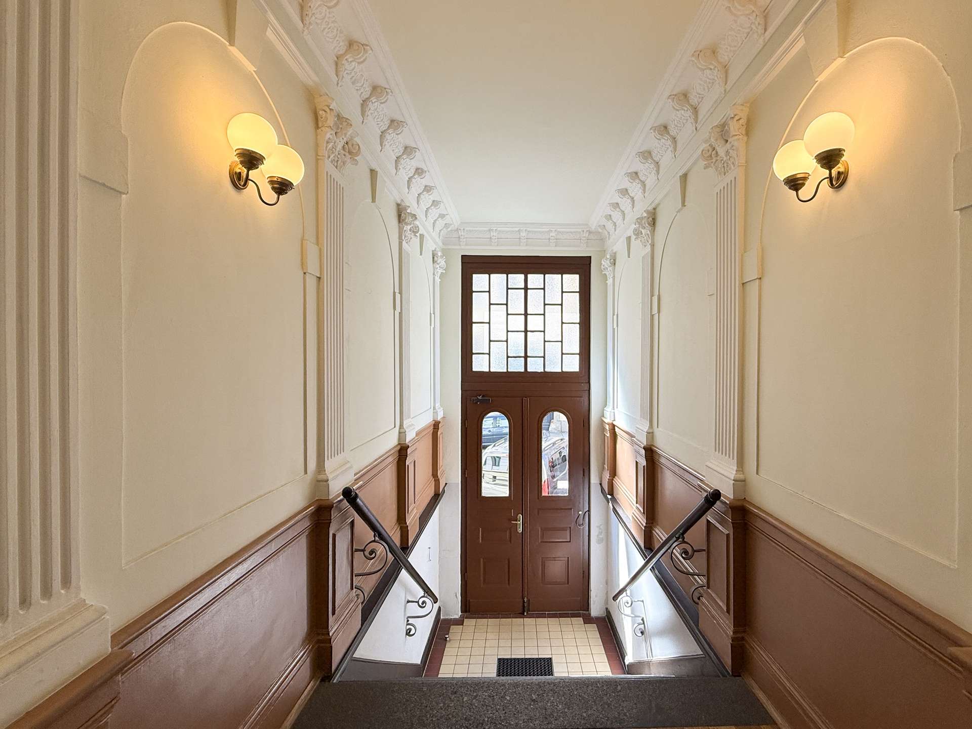 Elegant vintage hallway with ornate plaster molding, white walls, and warm wall lamps leading to a wooden door with frosted glass panes at the bottom of the stairs.