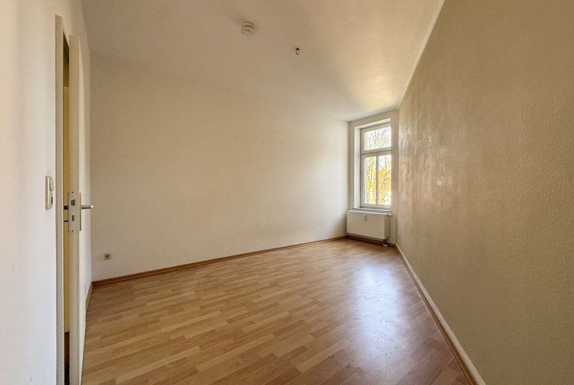 Empty sunlit room with wooden floor, beige walls, a window by the radiator, and a doorway on the left.