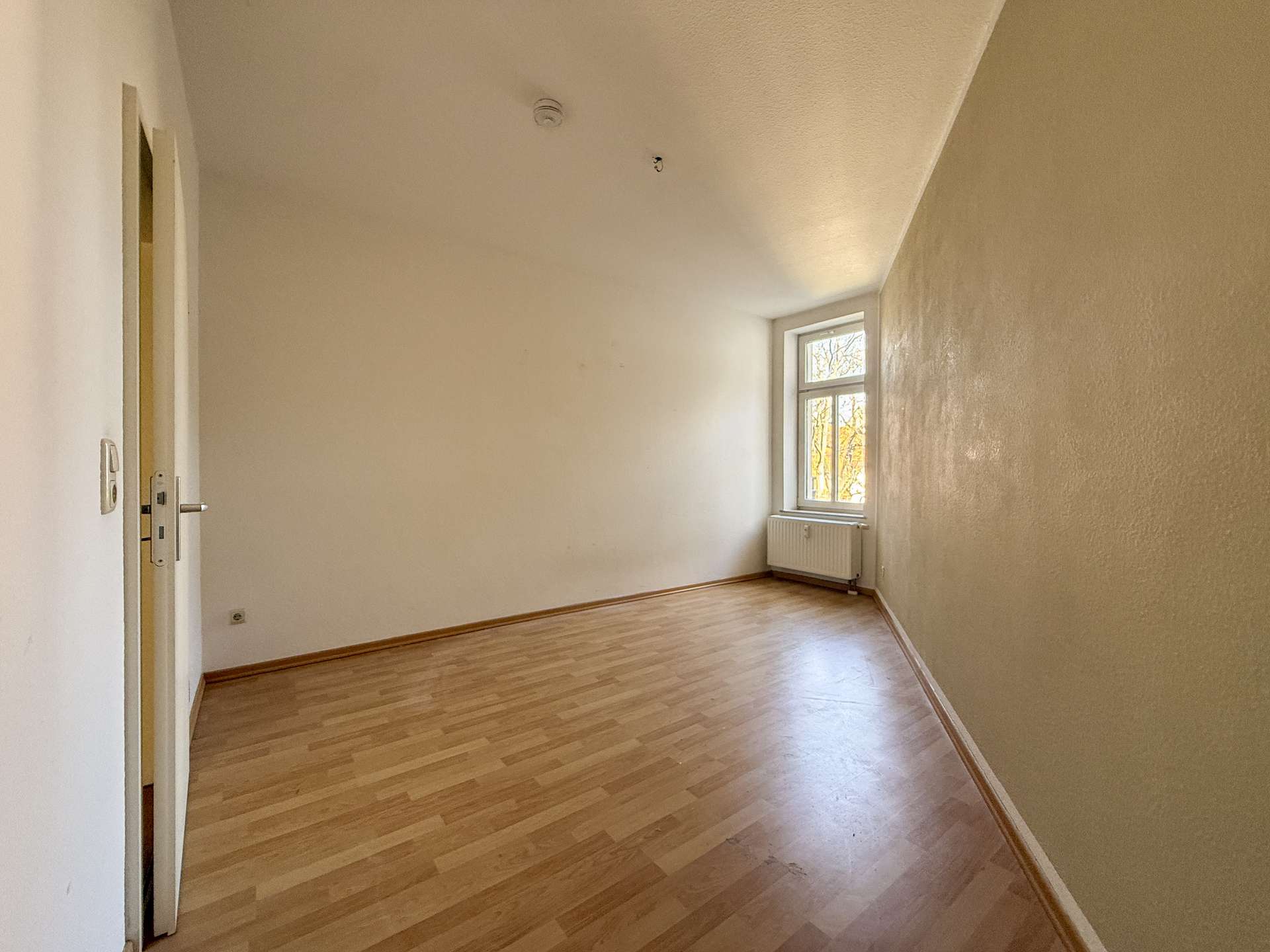 Empty sunlit room with wooden floor, beige walls, a window by the radiator, and a doorway on the left.