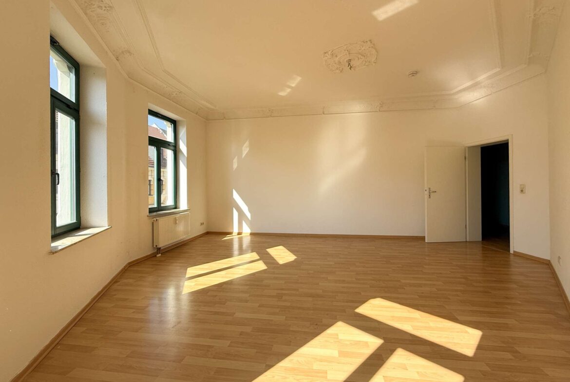 Sunlit empty room with a wooden floor, large green-framed windows, radiator, and an ornate ceiling moldings.