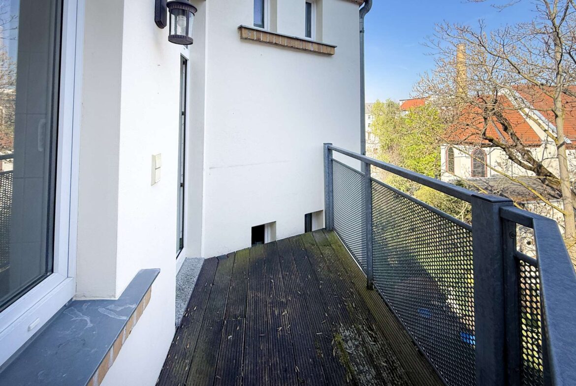 Wooden balcony along a white building with a metal mesh railing and a wall lantern by a window, view of trees and red rooftops in the distance.