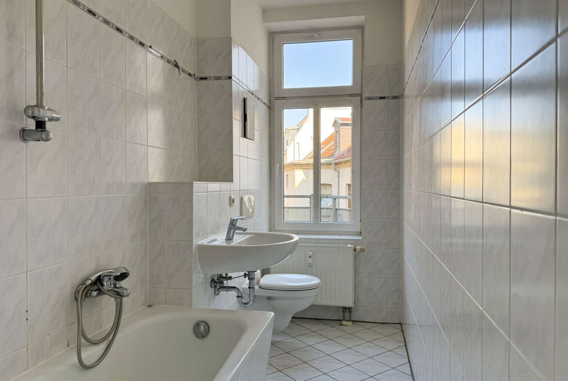 Narrow tiled bathroom with a bathtub along the left, a sink and toilet near a large window at the far end, letting in daylight the rooftops outside.