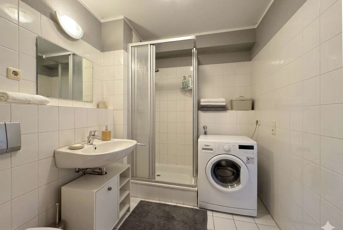 Modern bathroom with shower enclosure, washing machine, sink, and neatly folded towels on a shelf.
