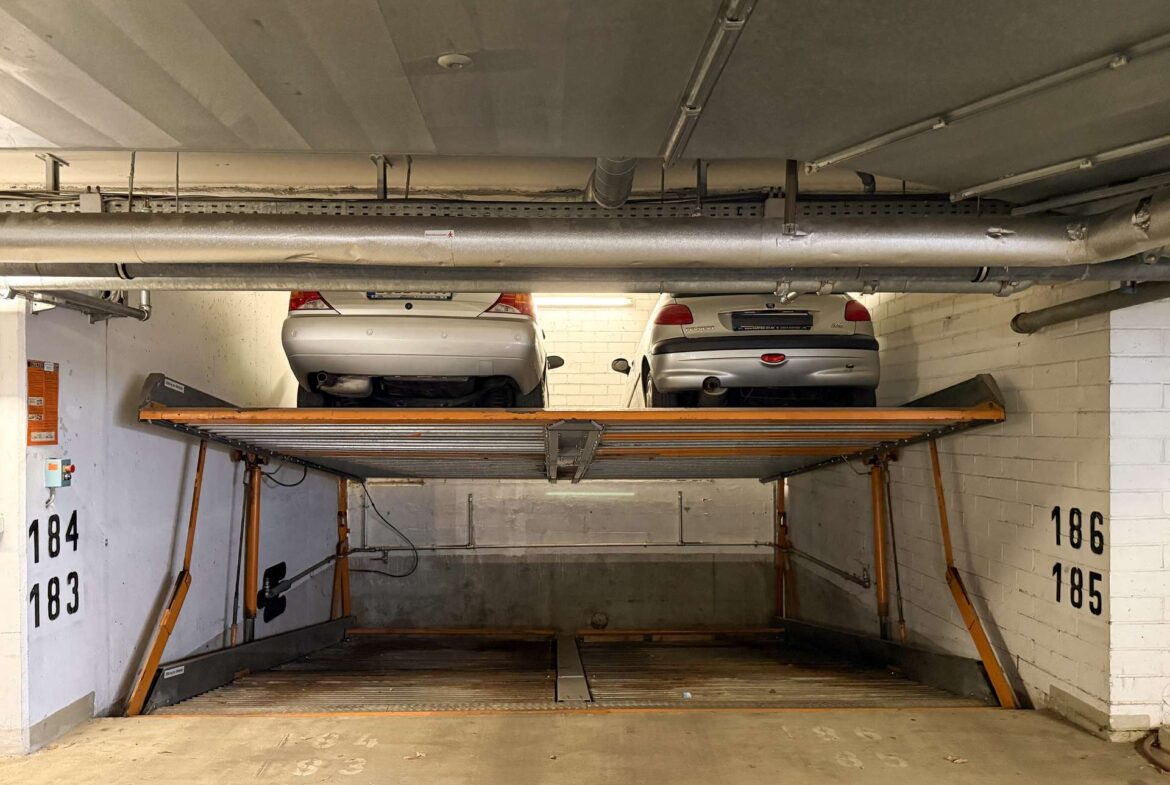 Underground parking garage with a two-car lift holding two silver sedans, elevated above the concrete floor. White walls show numbers 184–186, with exposed pipes and ceiling utilities overhead.