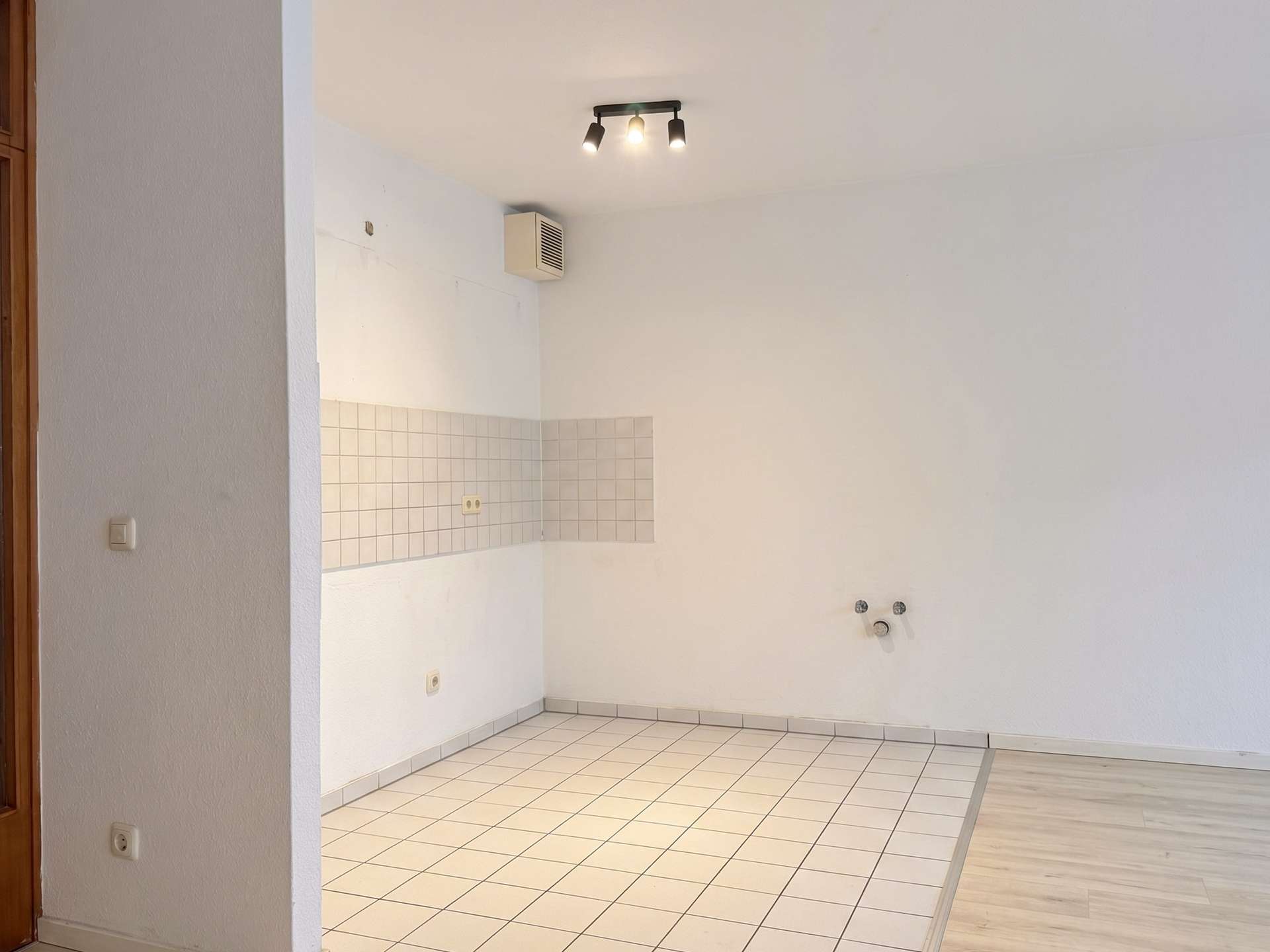 Empty kitchen corner with white tiled backsplash, outlets, and a three-bulb ceiling light; vent on the wall above.