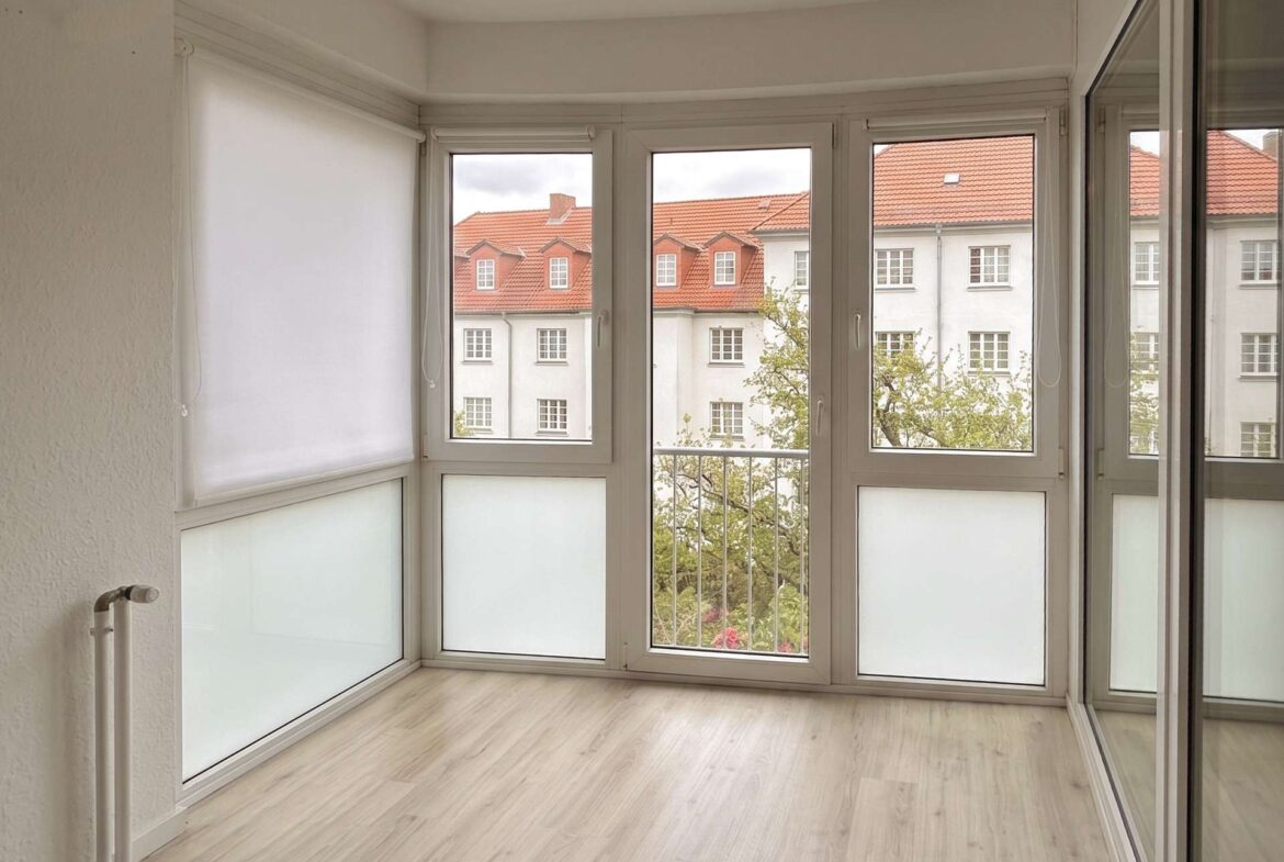 Bright sunroom with large white-framed windows, a left roller blind, and a view of red-tiled rooftops outside. A radiator is visible on the left wall.