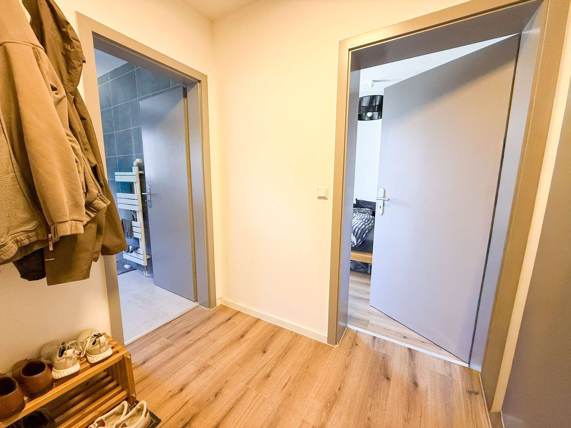 Hallway with a coat on the left and a shoe bench; two open doors reveal a blue-tiled bathroom on the left and a bedroom on the right.