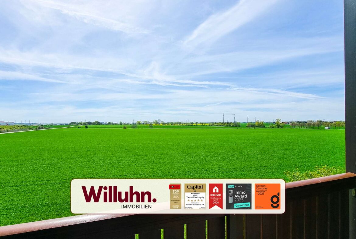 Wide rural landscape with a green field, blue sky, and a Willuhn Immobilien signboard in the foreground.