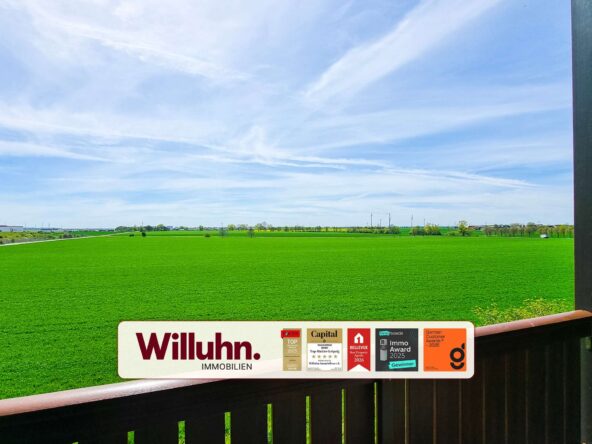 Wide rural landscape with a green field, blue sky, and a Willuhn Immobilien signboard in the foreground.
