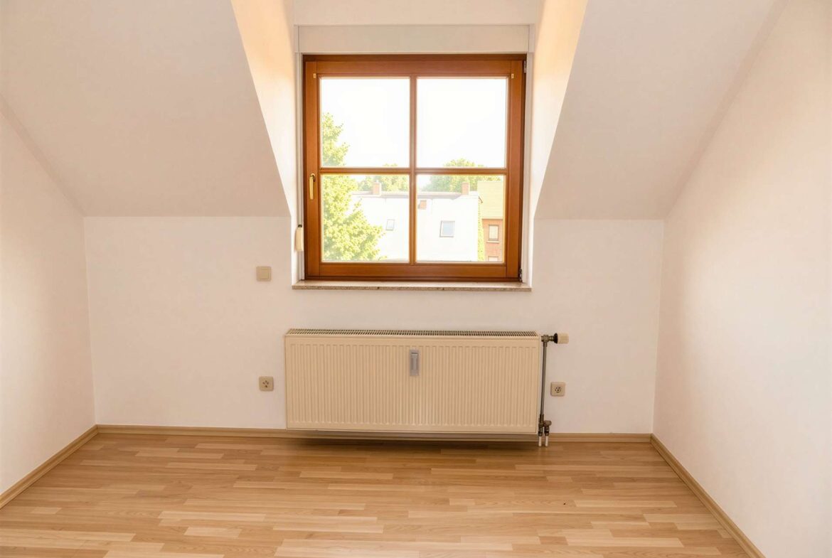 Empty attic room with a wooden-framed window, radiator beneath, and light hardwood flooring.